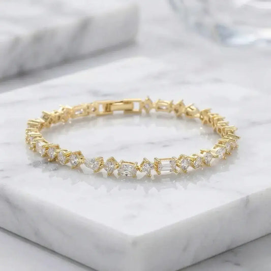 A gold-finish tennis bracelet featuring round, oval and teardrop zircon stones, displayed elegantly on a white marble surface.