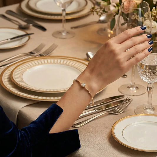 A model's arm resting on a sophisticated dinner table, featuring the Elegantia gold moissanite bangle, demonstrating its versatility for formal evening occasions.
