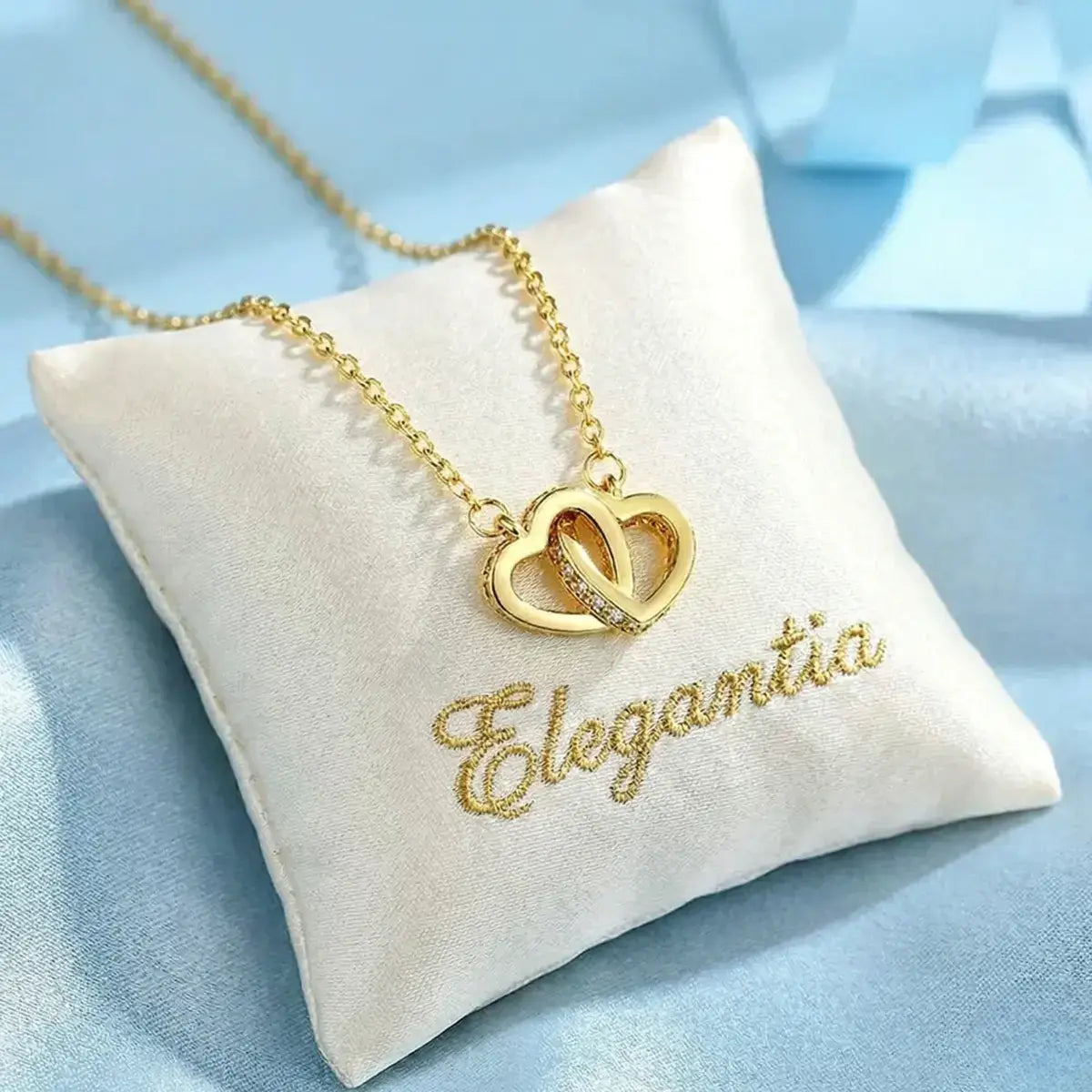 Small gold heart necklace resting on a white silk cushion with the word 'Elegantia' personalised in gold embroidery.