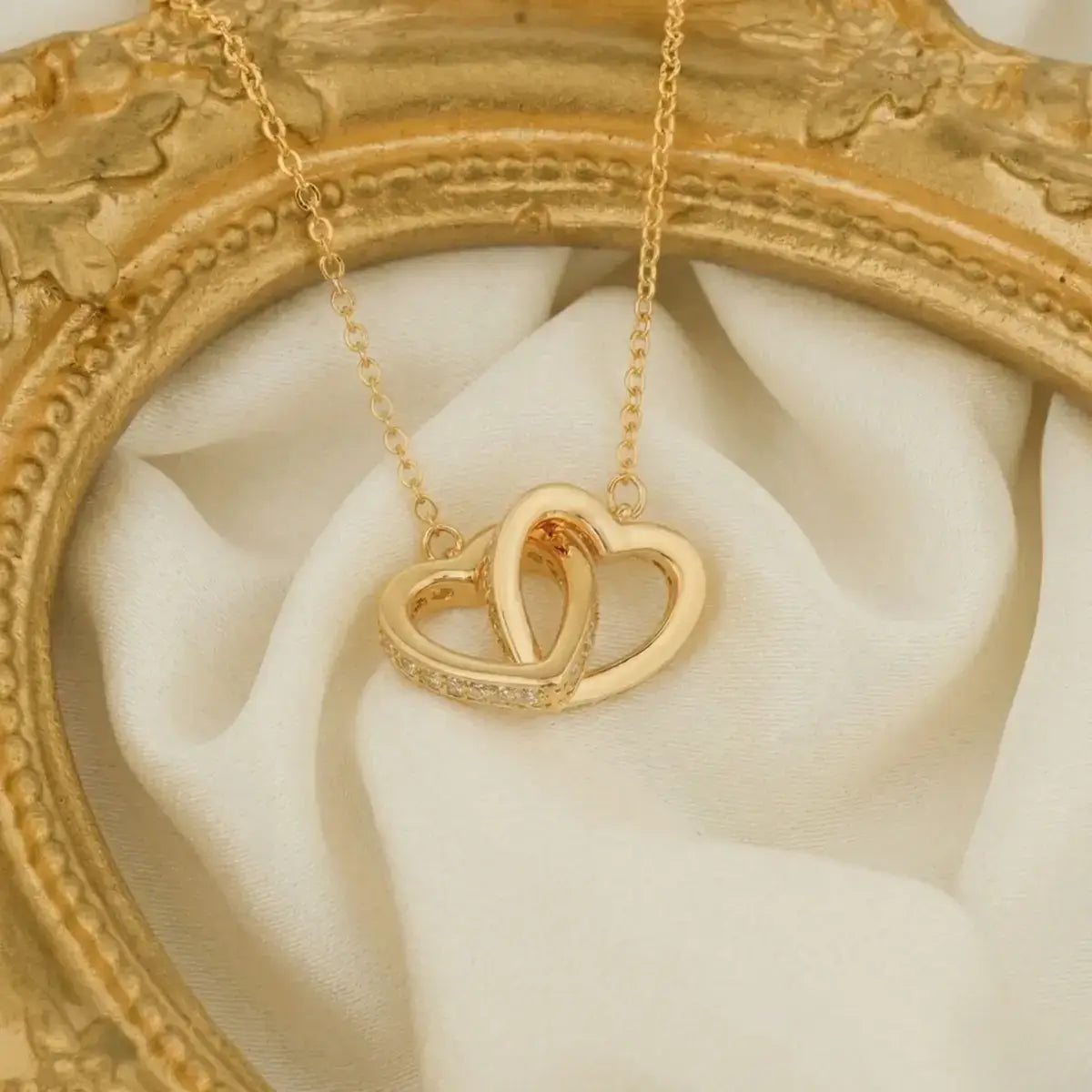 Gold necklace with interlocked heart pendant displayed on off-white draped fabric inside a vintage gold-coloured frame.
