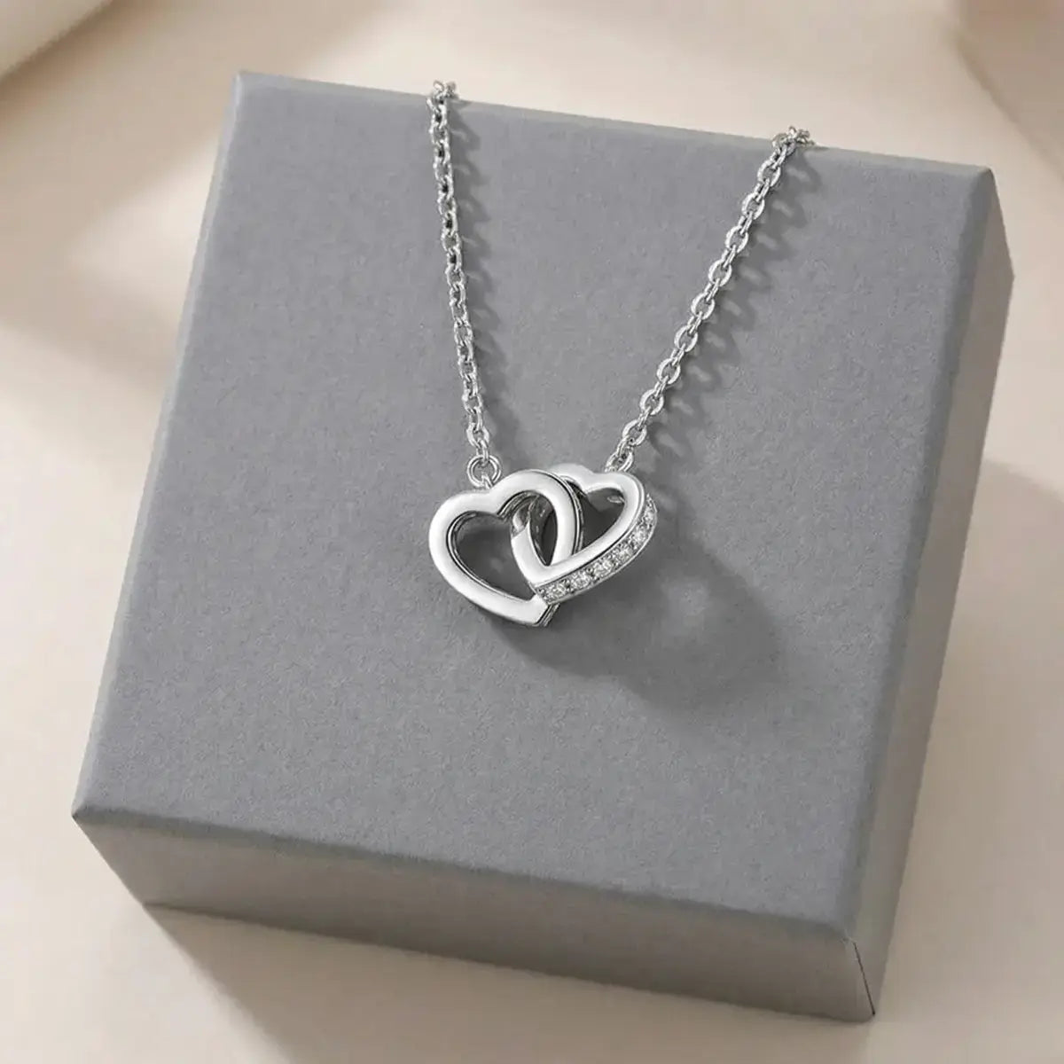 The Moissanite Interlocking Heart Necklace in sterling silver displayed on a minimalist grey jewellery box.